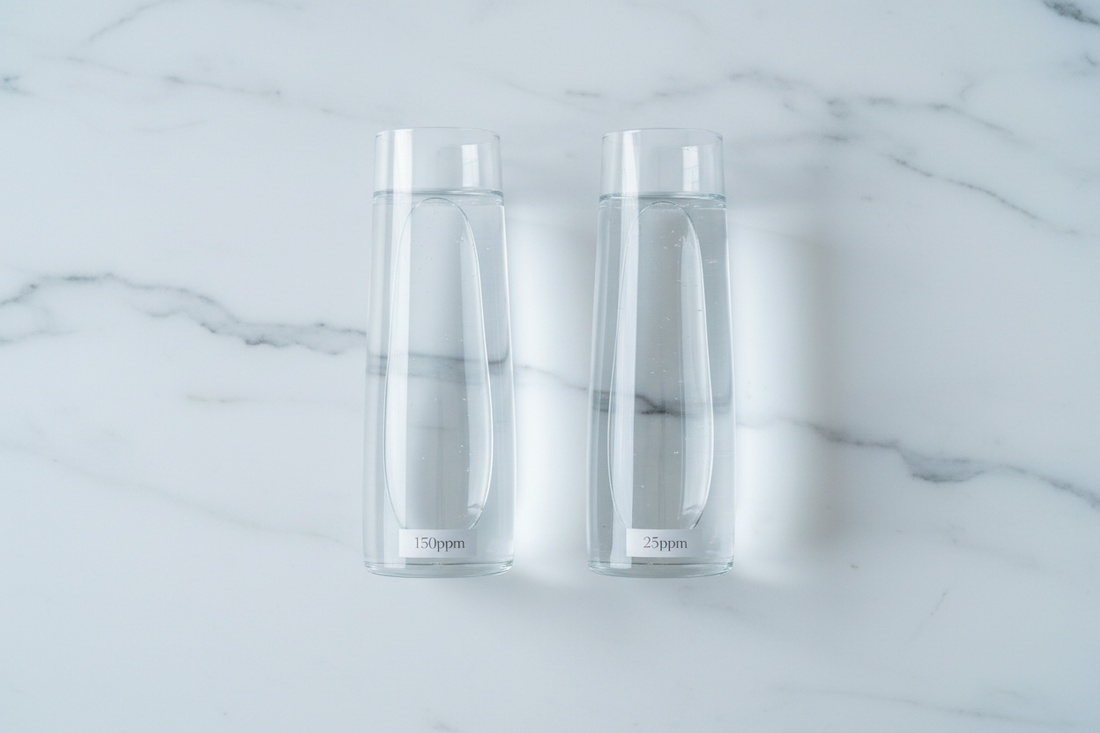 Two identical glass vessels of water side by side on a white marble surface, comparing regular water at 150ppm deuterium with Yavelle deuterium-depleted water at 25ppm