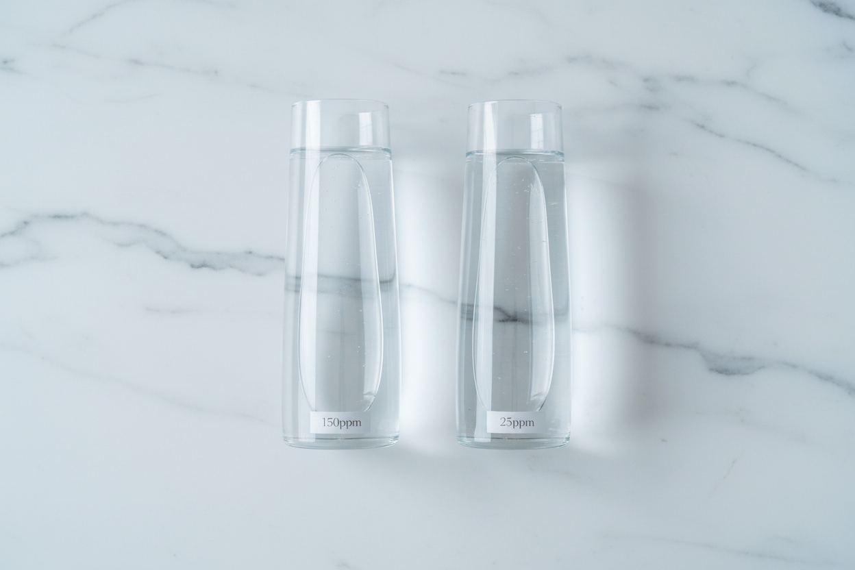 Two identical glass vessels of water side by side on a white marble surface, comparing regular water at 150ppm deuterium with Yavelle deuterium-depleted water at 25ppm